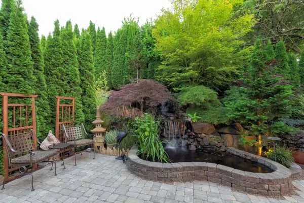 Serene outdoor landscape featuring a stone patio, lush greenery, a tranquil pond, and a waterfall, showcasing Laughter Family Hardscapes' landscape design expertise in creating peaceful outdoor spaces.