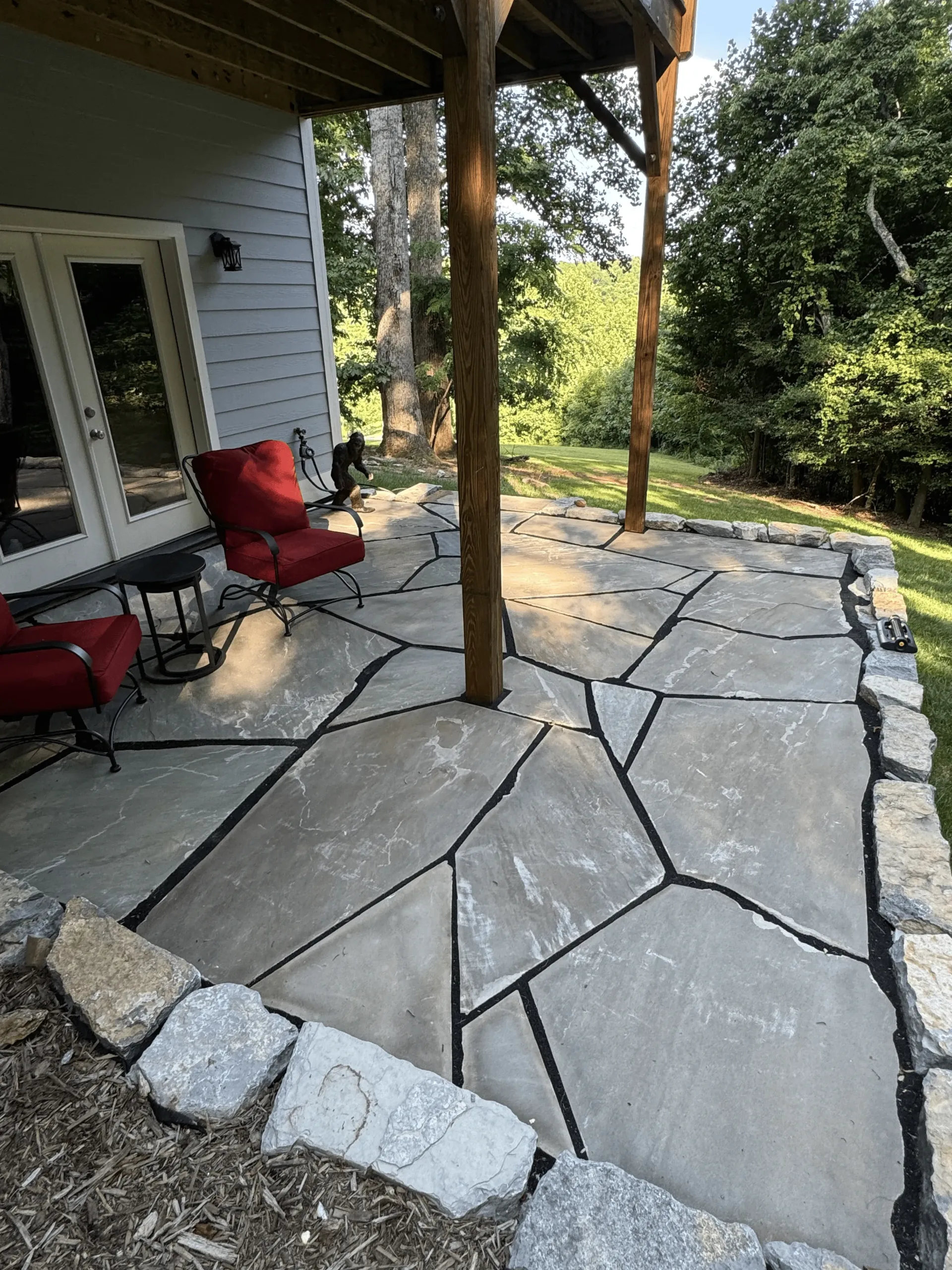 Patio area with irregular stone pavers, surrounded by natural landscaping, featuring red outdoor chairs and a wooden support beam, showcasing Laughter Family Hardscapes' hardscaping expertise in creating inviting outdoor spaces.