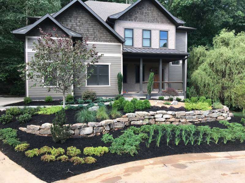 Front yard with stunning masonry work in Etowah, NC, showcasing a natural stone retaining wall surrounded by vibrant landscaping, including carefully arranged shrubs, perennials, and fresh mulch, enhancing the beauty of the modern two-story home.