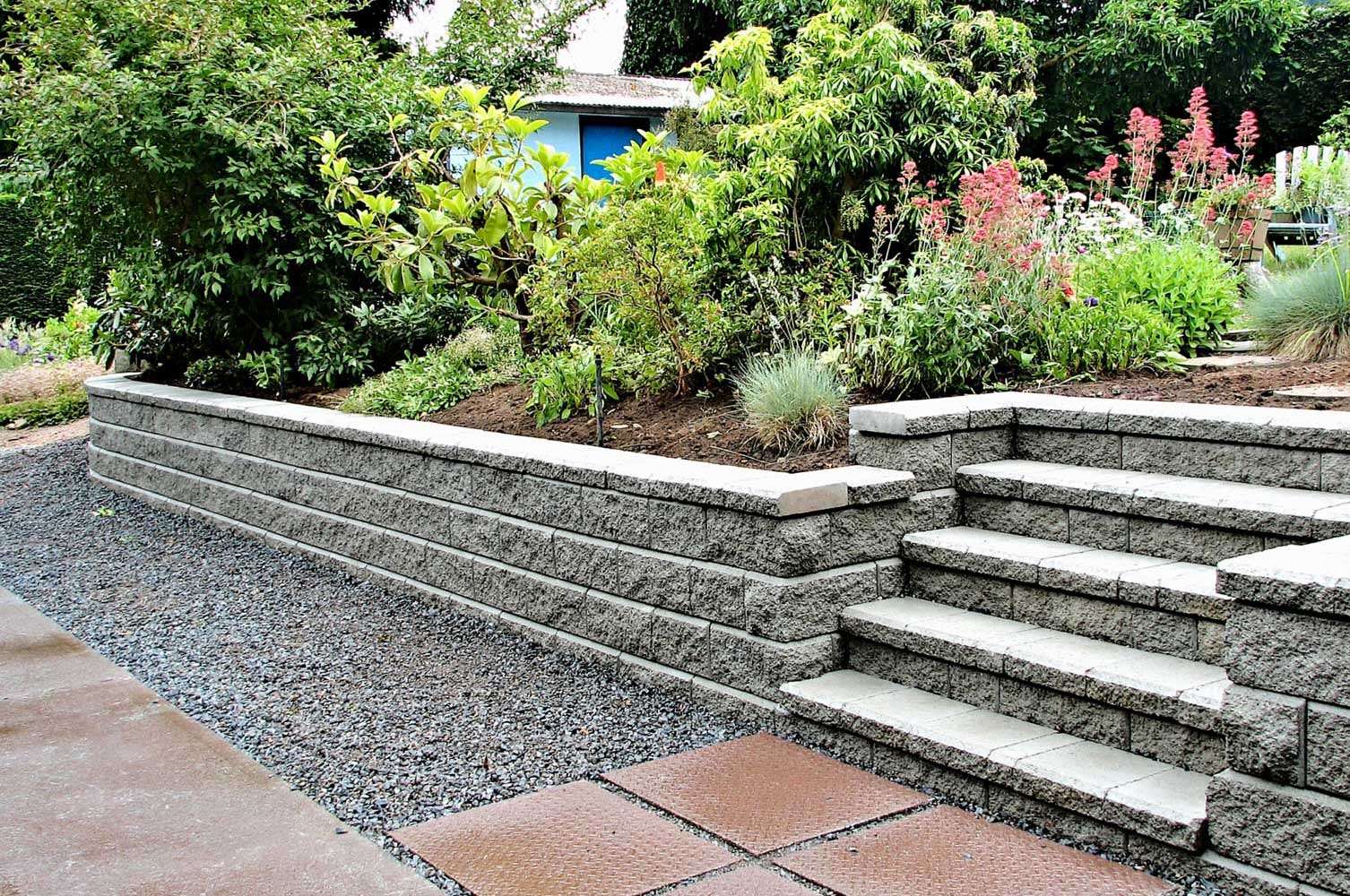 Stone retaining wall with built-in steps, beautifully bordered by lush greenery and vibrant flowers, highlighting expert masonry services in Etowah, NC.