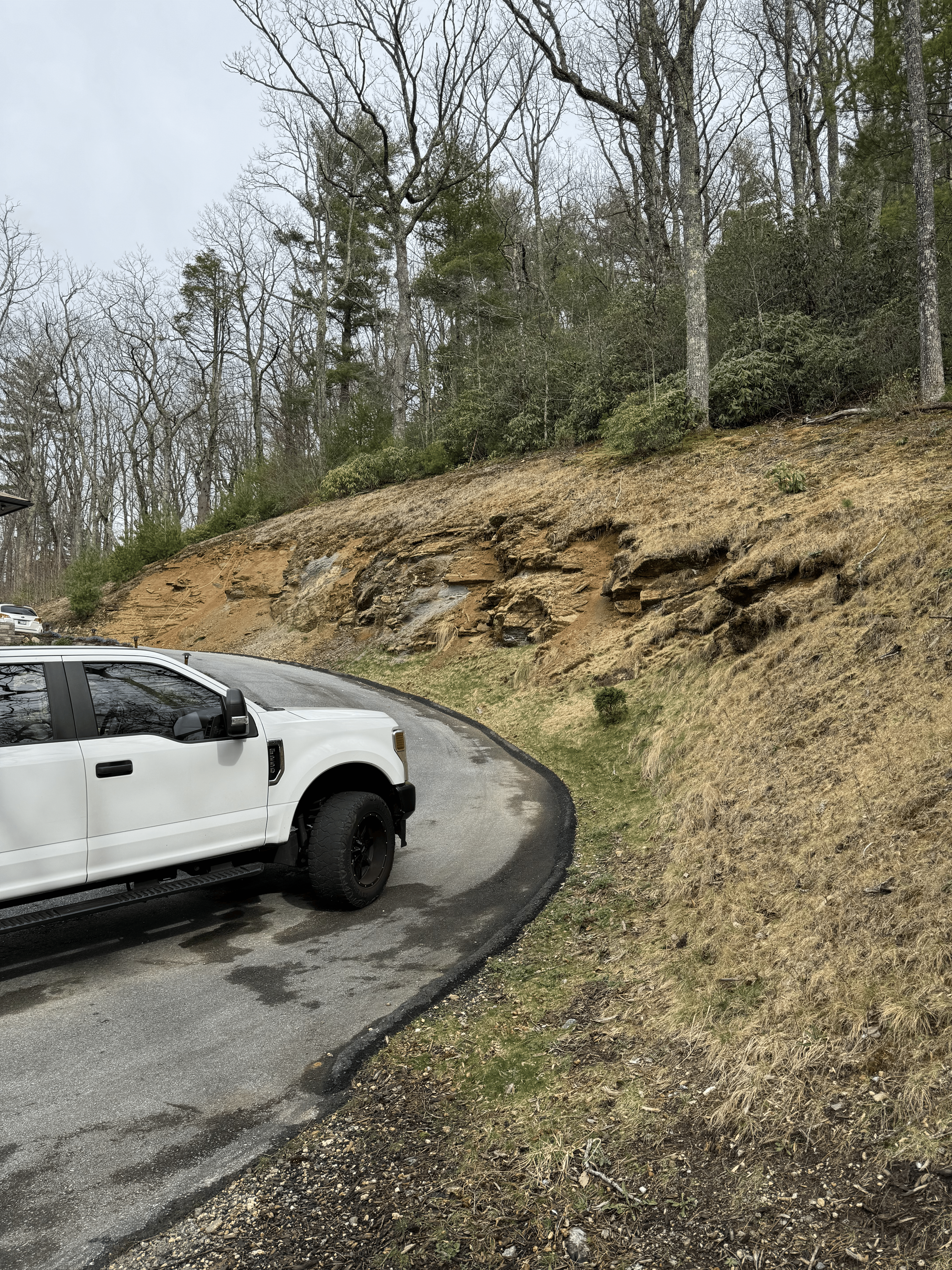White truck on winding road next to rocky hillside and wooded area, illustrating potential landscaping project site for Laughter Family Hardscapes.