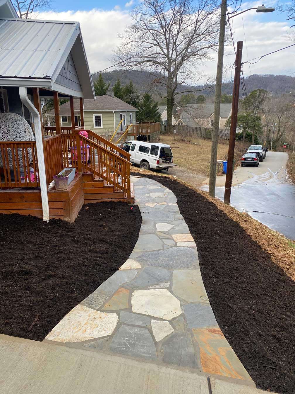 A beautifully crafted stone walkway curves gracefully alongside a wooden porch, bordered by freshly mulched landscaping, showcasing expert walkway installation in Etowah, NC.