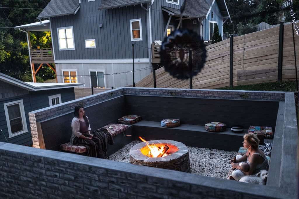 Cozy outdoor fire pit area with two women seated on benches, surrounded by landscaped walls and decorative cushions, showcasing a relaxing hardscaping design by Laughter Family Hardscapes in Asheville, NC.