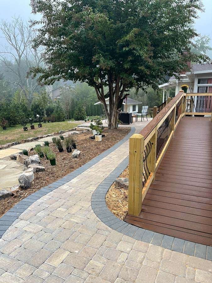 Curved stone pathway leading to a wooden deck, surrounded by landscaped greenery and a tree, showcasing hardscaping design by Laughter Family Hardscapes in Asheville, NC.