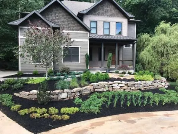 Beautifully landscaped home exterior featuring a mix of shrubs, ornamental trees, and stone retaining walls, emphasizing professional landscaping and hardscaping services by Laughter Family Hardscapes in Etowah, NC.