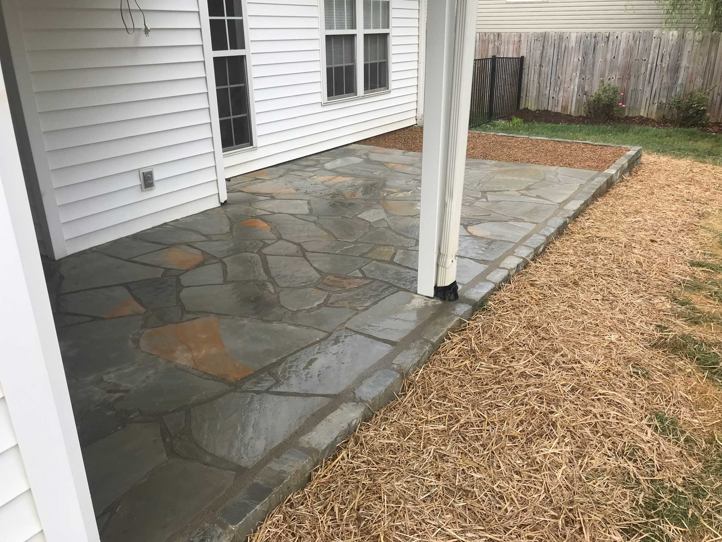 A freshly installed flagstone patio extends from a white residential home, bordered by straw-covered soil and mulch, demonstrating professional hardscaping services in Etowah, NC.