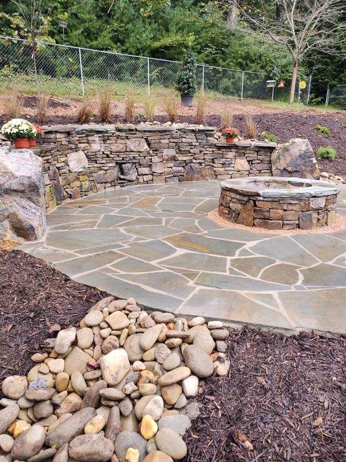 Custom hardscaping design featuring a stone seating area, circular fire pit, and natural stone pathway, surrounded by landscaped greenery and decorative plants, showcasing Laughter Family Hardscapes' craftsmanship in outdoor space transformation in Asheville, NC.