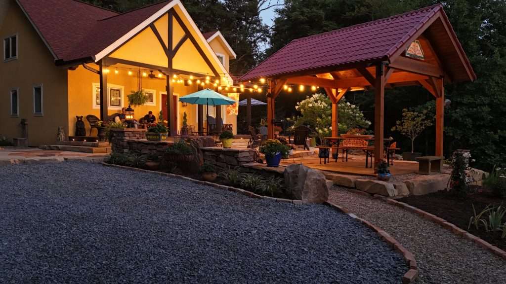 Outdoor kitchen with illuminated patio area, featuring a wooden gazebo, dining table, and bright umbrella, surrounded by lush greenery and decorative plants, enhancing an inviting outdoor living space.