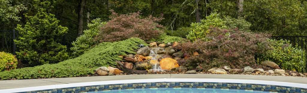 Custom landscaping with a stone waterfall feature and lush greenery near a pool, showcasing Laughter Family Hardscapes' design expertise in Asheville, NC.