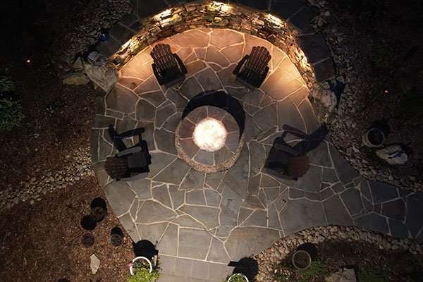 Outdoor hardscaping design featuring a circular stone fire pit surrounded by wooden chairs, illuminated at night, showcasing Laughter Family Hardscapes' custom landscaping work in Asheville, NC.