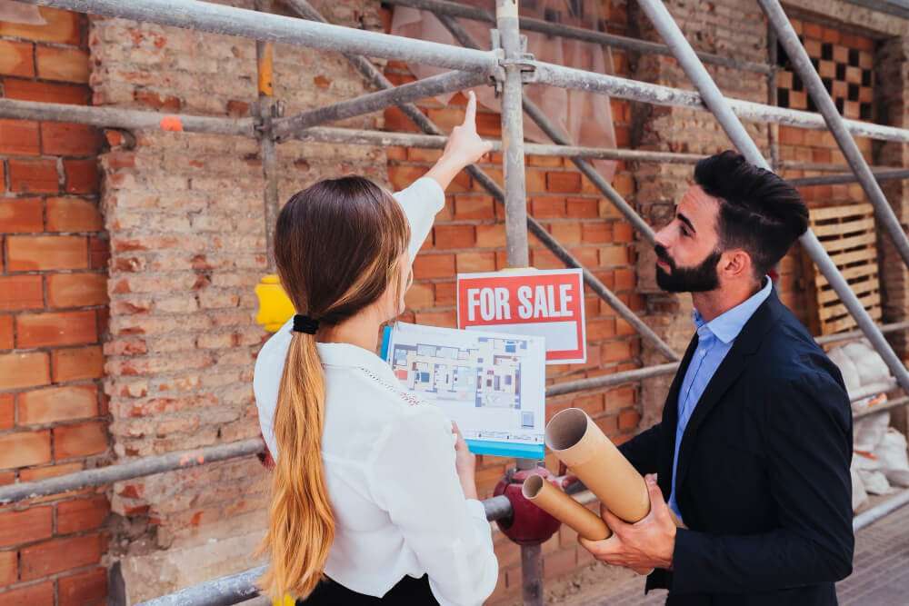 Real estate agents discussing property plans in front of a "For Sale" sign and scaffolding, emphasizing outdoor space potential for hardscaping projects.