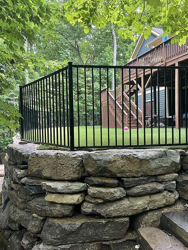 Black metal fence enclosing a landscaped yard with green grass and a pink ball, adjacent to a wooden deck and surrounded by trees, showcasing a hardscaping project by Laughter Family Hardscapes in Asheville, NC.
