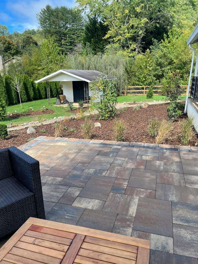 Patio area featuring stone pavers, wooden table, and black wicker chair, surrounded by landscaped garden with shrubs and small trees, showcasing Laughter Family Hardscapes' custom outdoor design in Asheville, NC.