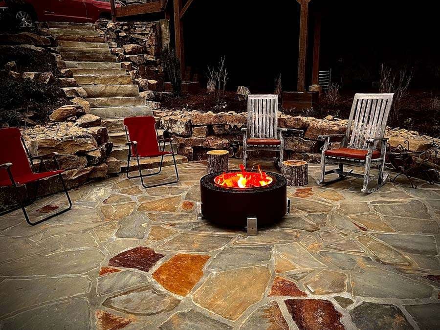 Fire pit surrounded by stone patio, red and white chairs, and rustic wooden stools in a cozy outdoor setting, showcasing hardscaping features for enhancing outdoor living spaces in Etowah, NC.