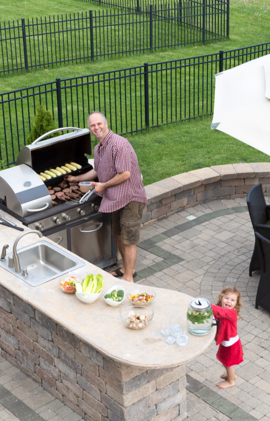 Man grilling burgers and vegetables in a custom-built outdoor kitchen with a stone countertop and built-in sink, showcasing professional outdoor kitchen services in Etowah, NC."