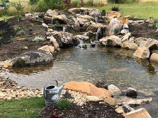 Water feature with cascading rocks and a tranquil pond, surrounded by natural landscaping and a metal watering can, showcasing Laughter Family Hardscapes' custom outdoor design in Asheville, NC.