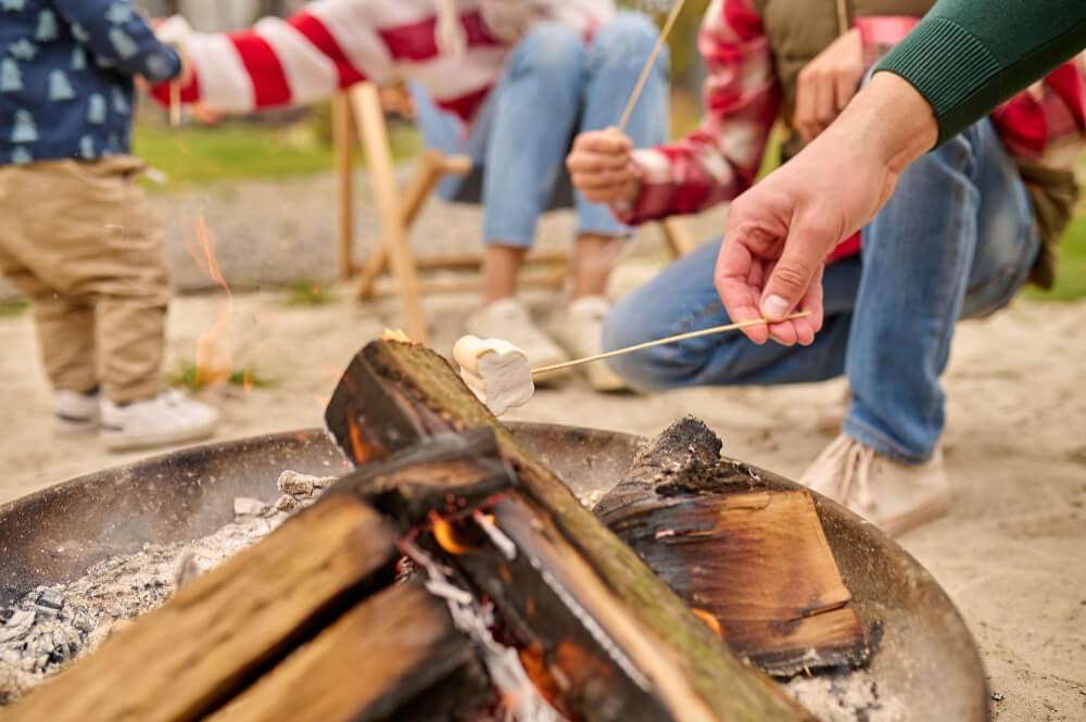 Male hand holding marshmallow on skewer over outdoor bonfire, with family members in background enjoying DIY fire pit experience.