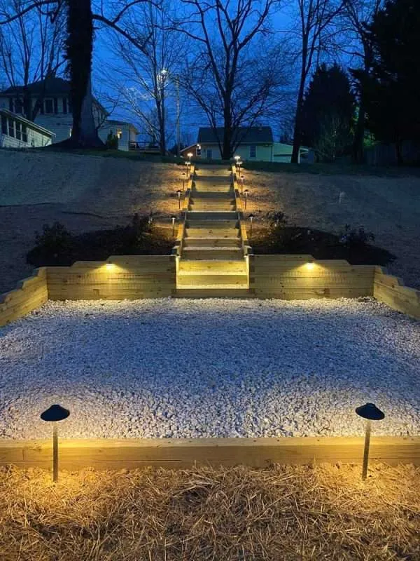 Illuminated wooden steps leading to a landscaped area with gravel, surrounded by soft lighting, showcasing a hardscaping design by Laughter Family Hardscapes in Etowah, NC.
