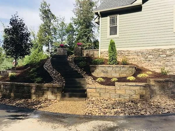 Landscaped outdoor space featuring stone retaining walls, a staircase leading to a house, and colorful plants, showcasing professional hardscaping design by Laughter Family Hardscapes in Etowah, NC.