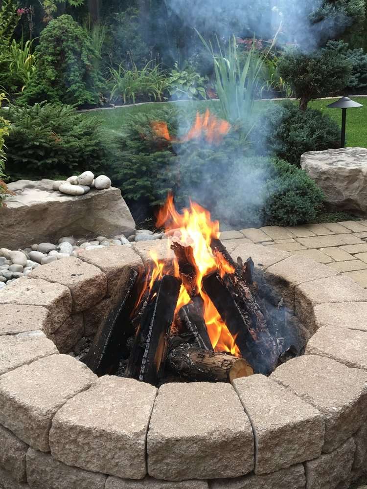 Fire pit with burning logs surrounded by stone seating, set in a lush garden landscape, enhancing outdoor living space ambiance.