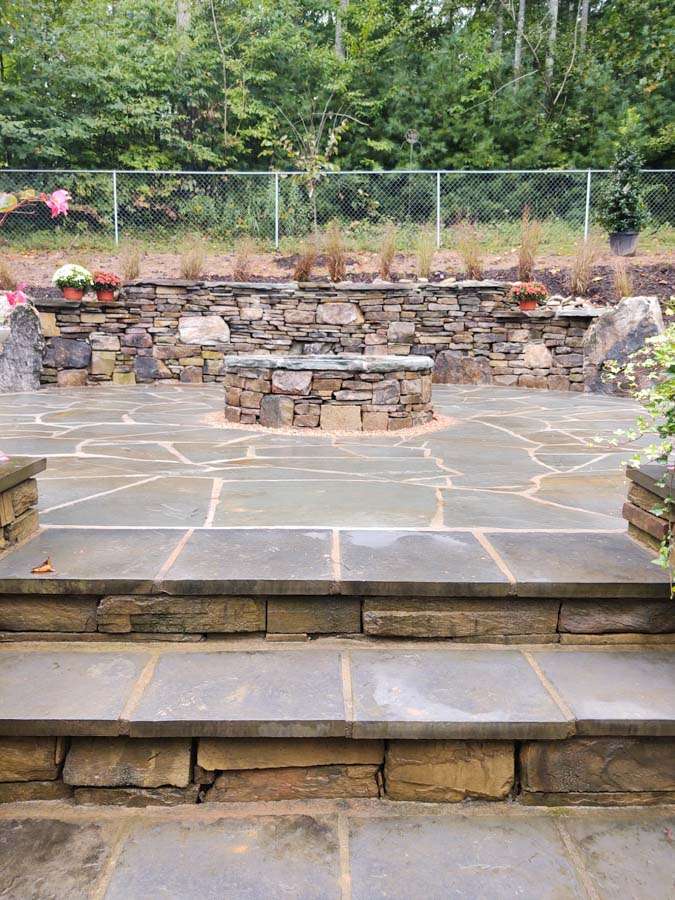 Hardscaped outdoor space featuring a stone fire pit surrounded by a circular stone wall, flagstone patio, and lush greenery in the background, showcasing Laughter Family Hardscapes' custom landscaping services in Asheville, NC.