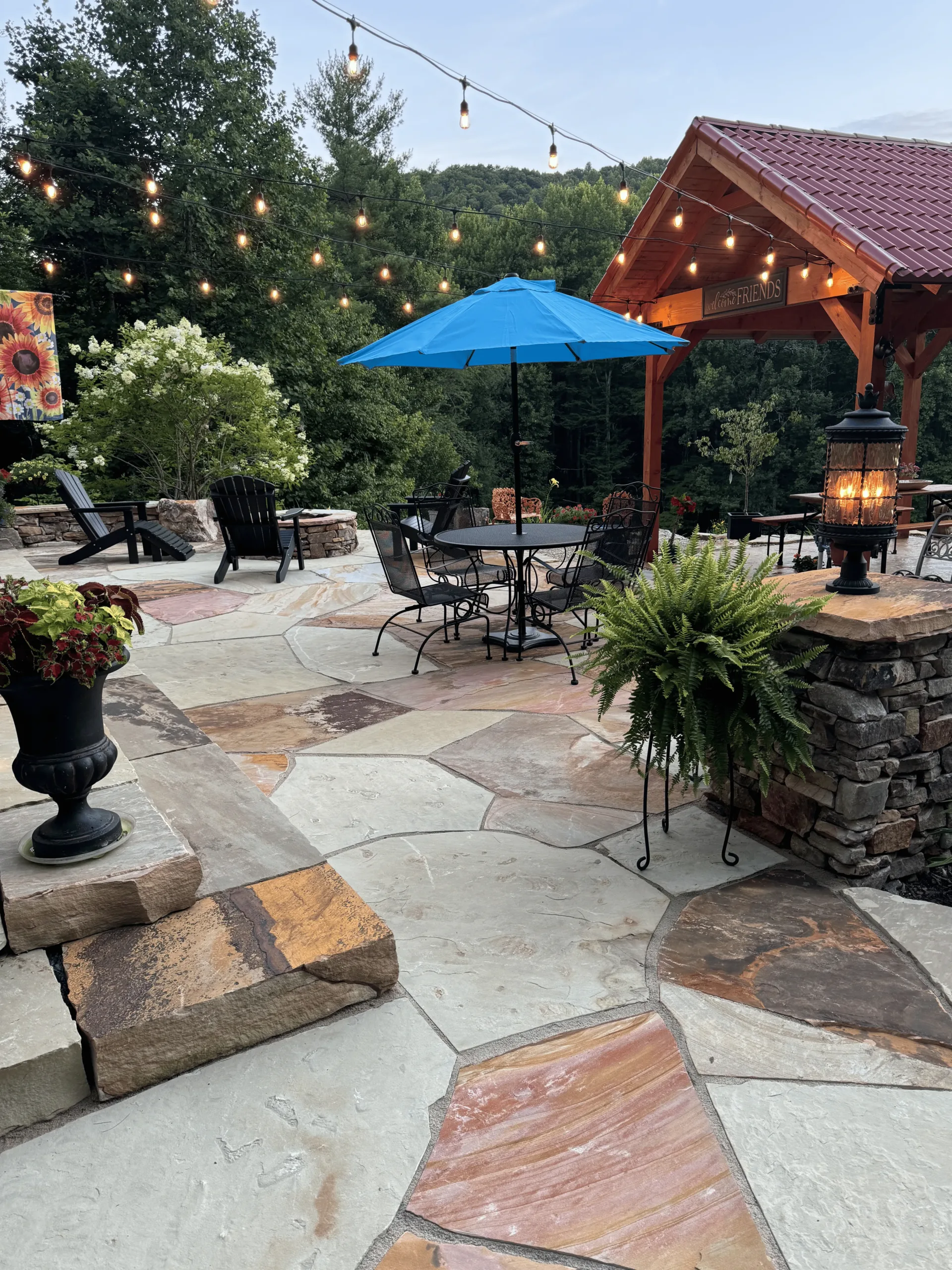 Outdoor patio featuring natural stone flooring, a blue umbrella over a dining table, wooden Adirondack chairs, decorative lantern, and string lights, showcasing a beautifully designed landscape for Laughter Family Hardscapes in Etowah, NC.
