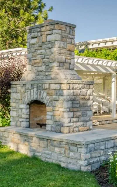 Stone outdoor fireplace with a rustic design, surrounded by lush greenery and a patio area, emphasizing hardscaping and outdoor living spaces by Laughter Family Hardscapes.