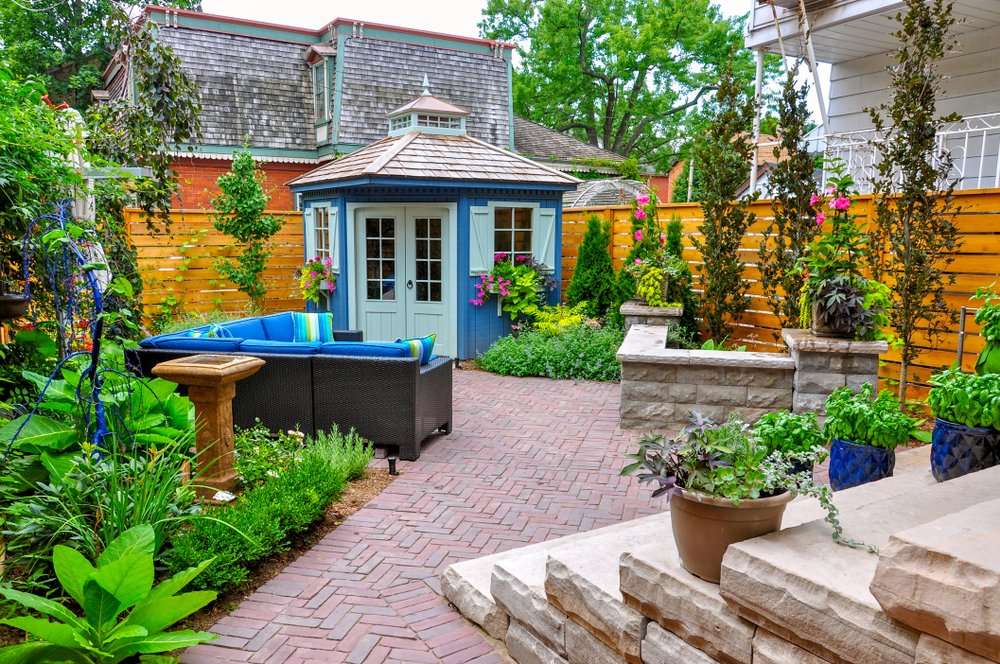Cozy backyard hardscaping design featuring a blue garden shed, a comfortable seating area with vibrant cushions, and lush greenery, showcasing pathways and planters for an inviting outdoor living space.