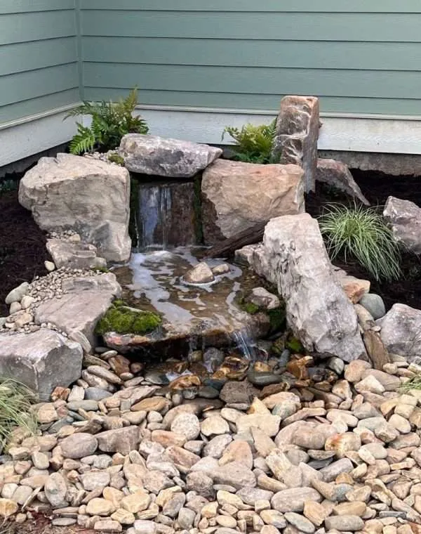 Water feature with cascading waterfall and natural stones, surrounded by decorative rocks and greenery, showcasing landscaping design by Laughter Family Hardscapes.