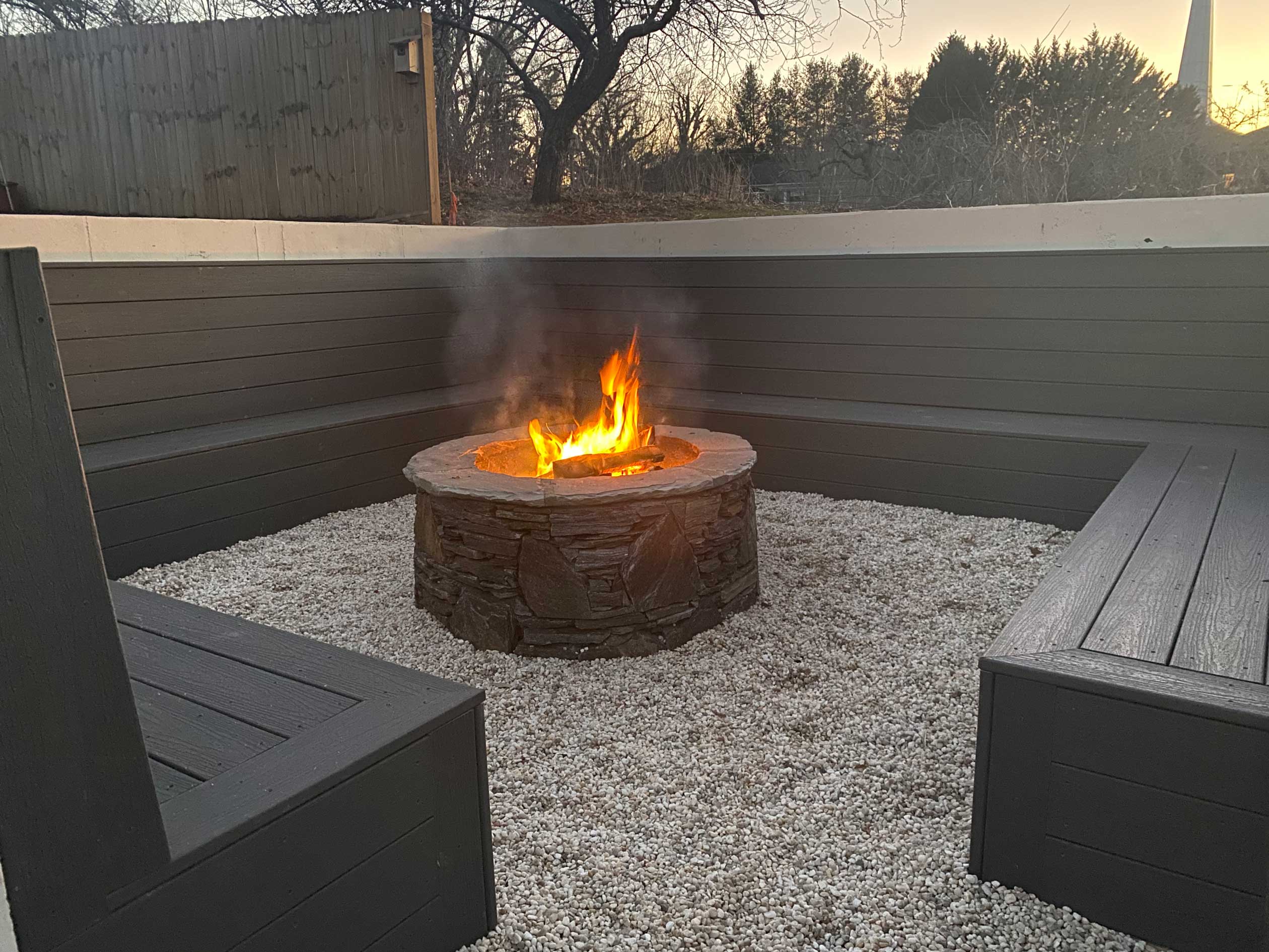 A cozy outdoor seating area with a central stone fire pit surrounded by custom-built wooden benches, perfect for evening gatherings, highlighting professional fire pit installation in Etowah, NC.