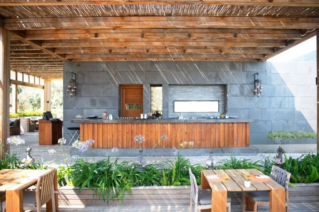 Custom outdoor kitchen with stone island, wooden accents, and bar-style seating, surrounded by lush greenery and patio dining area, enhancing outdoor entertaining space.