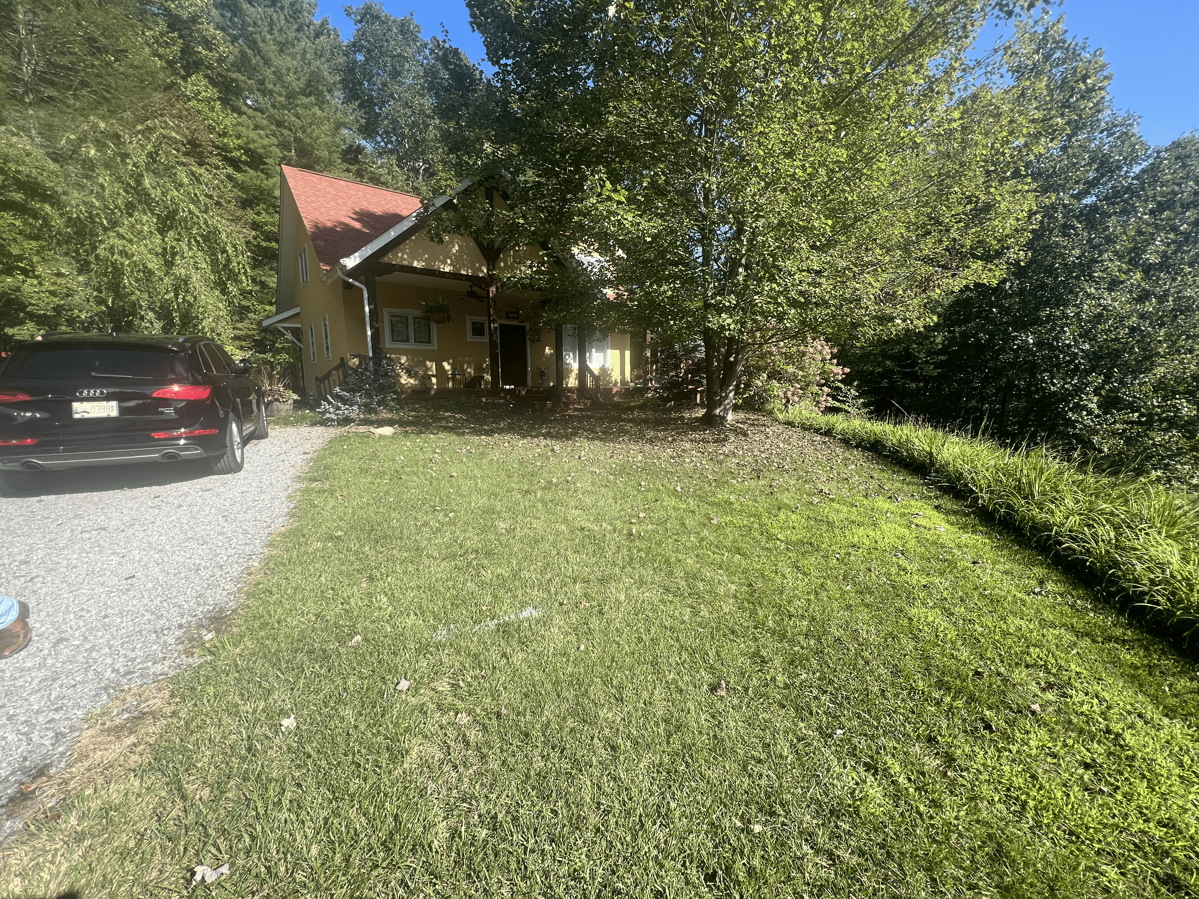 Residential landscaping featuring a yellow house with a red roof, surrounded by trees and a manicured lawn, with a black car parked on a gravel driveway, showcasing potential for landscaping enhancement.