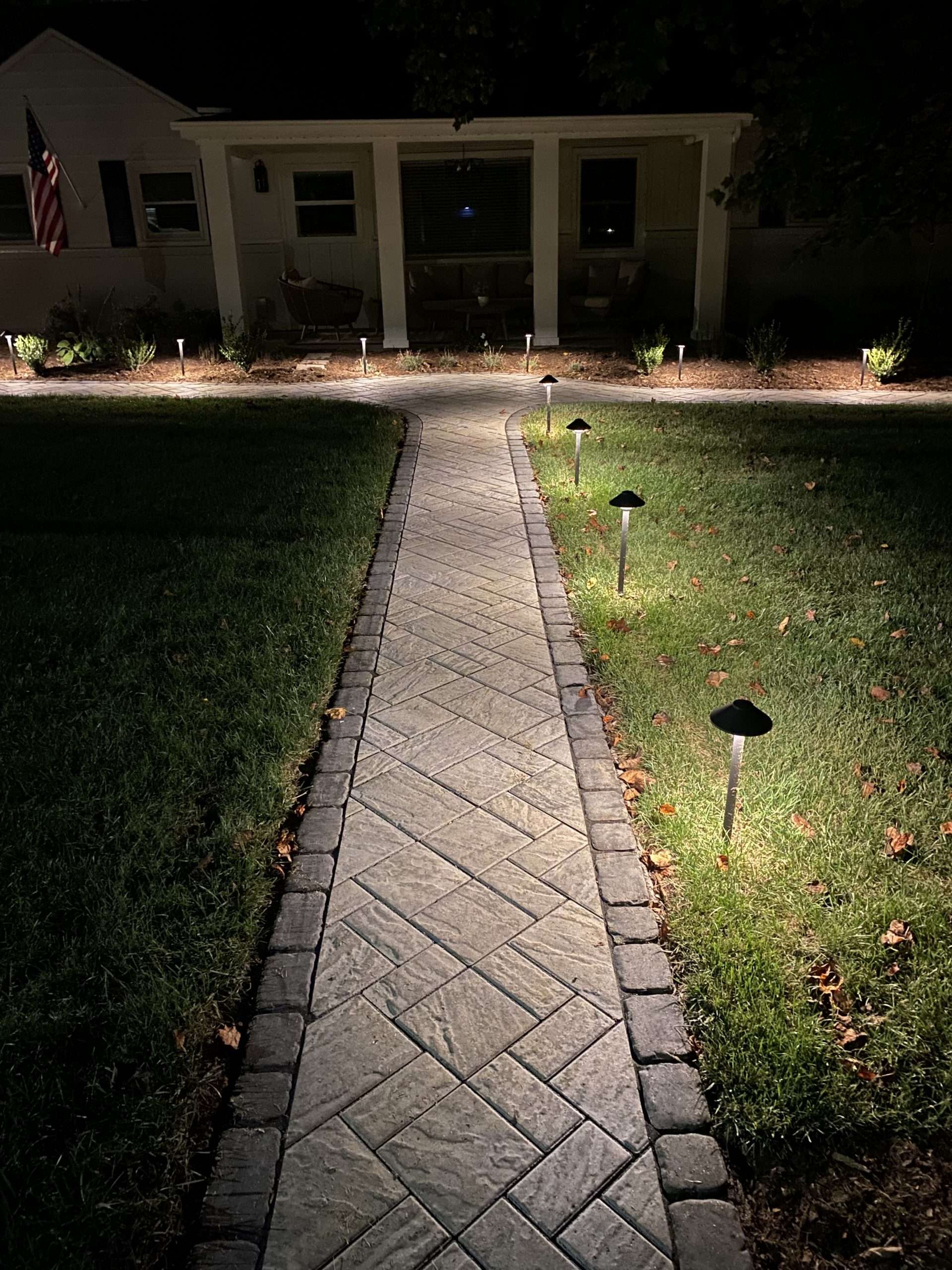 Illuminated walkway with patterned stone path leading to a home, surrounded by soft landscape lighting and grass, showcasing Laughter Family Hardscapes' custom hardscaping design in Asheville, NC.